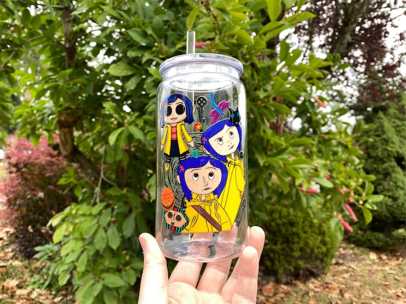 May include: A clear glass tumbler with a lid and straw, decorated with a colourful cartoon graphic. The design features three characters in yellow raincoats and blue hair. The background is a lush green with foliage.