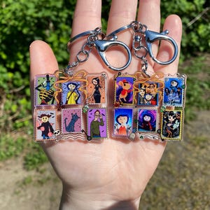 May include: Two keychains with silver-colored clasps and chains. Each keychain holds a collection of small, rectangular acrylic charms featuring colorful illustrations of characters. The charms are arranged in a grid-like pattern, with a variety of images.