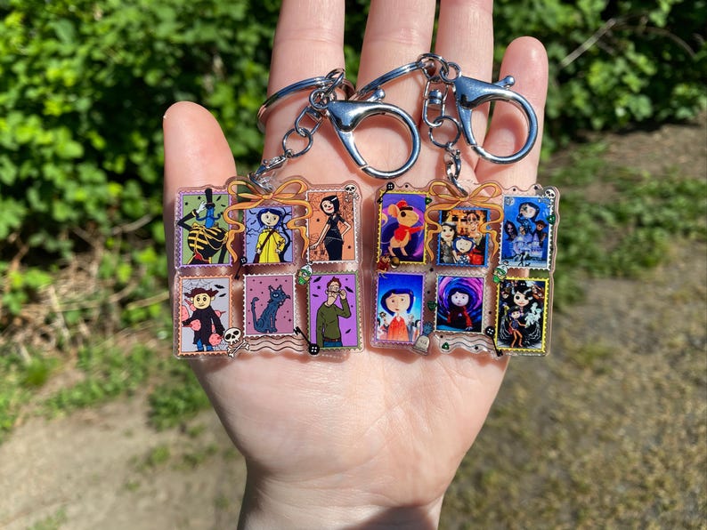 May include: Two keychains with silver-colored clasps and chains. Each keychain holds a collection of small, rectangular acrylic charms featuring colorful illustrations of characters. The charms are arranged in a grid-like pattern, with a variety of images.