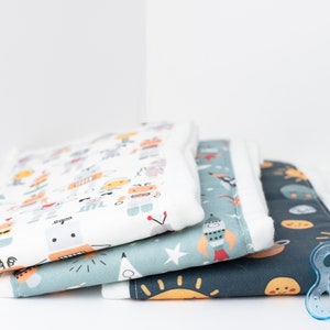 May include: Three baby burp cloths with different colorful patterns. The top cloth has a white background with a repeating pattern of robots, stars, and other fun designs. The middle cloth has a blue background with a repeating pattern of stars, rockets, and planets. The bottom cloth has a dark blue background with a repeating pattern of suns, moons, and stars. A blue pacifier with a hedgehog design is in the foreground.