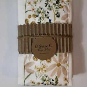 May include: Two white burp cloths with a floral pattern in shades of brown and green. The cloths are packaged together with a brown cardboard band and a label that reads "O Grace C. Burp Cloths, Huntsville, AL".