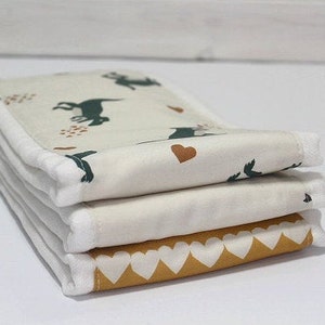 May include: A stack of three white fabric burp cloths with a white trim. The top cloth has a green and brown animal print with a small heart. The middle cloth is plain white. The bottom cloth has a yellow and white heart pattern.