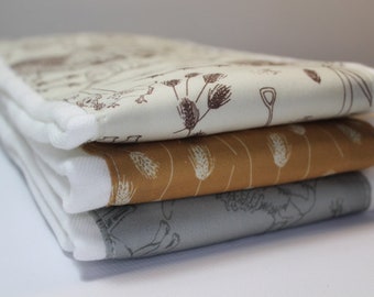 May include: Three fabric squares stacked on top of each other. The top square is white with a brown and white floral pattern. The middle square is brown with a white wheat pattern. The bottom square is gray with a brown and white floral pattern.