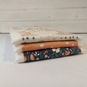May include: Three fabric squares stacked on top of each other. The top square is white with a gray and yellow rainbow, gray raindrops, and small white hearts. The middle square is peach with a white heart. The bottom square is dark blue with a white floral pattern.