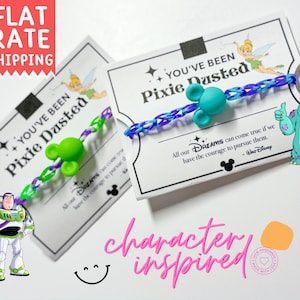 May include: Character-inspired bracelets with Mickey Mouse-shaped beads. One bracelet is green and purple, the other is blue and purple. The cards read "You've Been Pixie Dusted" with Disney characters. The words "character inspired" are in pink.
