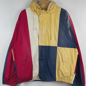 May include: A colour-blocked windbreaker jacket featuring a hood with drawstrings, a front zip closure, and two side pockets. The garment is constructed from panels of yellow, red, white, and navy blue fabric, with long sleeves and elastic cuffs.