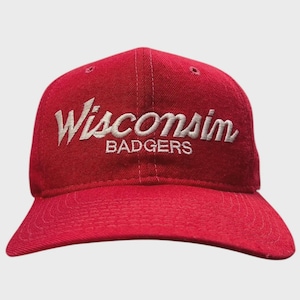 May include: A red baseball cap with the word "Wisconsin" embroidered in white script above the word "Badgers". The cap has a curved brim and a six-panel design.