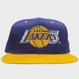 Può includere: Un cappellino da baseball viola con visiera gialla che presenta il logo ricamato dei Los Angeles Lakers sul davanti. Il logo è composto da un pallone da basket giallo con il testo viola Los Angeles Lakers e accenti bianchi. Sono visibili piccoli occhielli gialli.