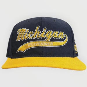 Może przedstawiać: Granatowa czapka z daszkiem z płaskim żółtym daszkiem. Z przodu znajduje się żółty haft z napisem Michigan kursywą oraz słowem WOLVERINES białymi literami na żółtym łuku znajdującym się pod głównym napisem.