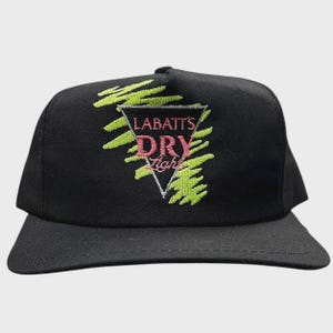 Könnte beinhalten: Schwarze Baseballkappe mit flachem Schirm. Die Kappe hat ein rosa-weißes Dreieckslogo mit den Worten "LABATT'S DRY Light" und grünen abstrakten Formen. Die Kappe ist aus strapazierfähigem Stoff gefertigt und hat einen klassischen, lässigen Stil.