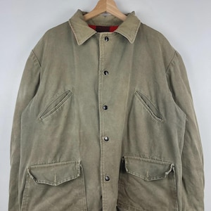 May include: A tan canvas jacket featuring a pointed collar, a front button closure with dark buttons, two slanted chest pockets, and two large flap pockets at the waist. The interior lining is visible at the hem and collar in a red plaid pattern.