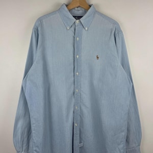 Puede incluir: Una camisa de manga larga en color azul claro con cuello abotonado y un pequeño logotipo bordado en el pecho izquierdo. La camisa está confeccionada en un tejido ligero e incluye una tapeta frontal completa con botones de color blanco.