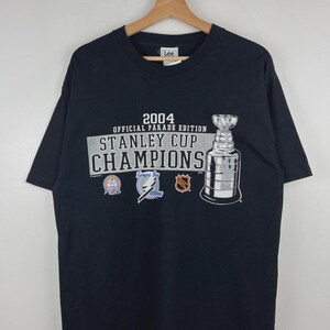 May include: A black short-sleeve t-shirt featuring a 2004 Official Parade Edition Stanley Cup Champions graphic. The design includes the Stanley Cup trophy, the Tampa Bay Lightning logo, and the NHL logo printed in white, blue, and orange ink.