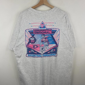 May include: Light grey t-shirt with a vibrant graphic promoting the Griffith Memorial Racquetball Tournament '95. The design incorporates a racquetball player, geometric patterns, and text in pink and blue.