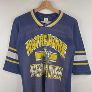 Może przedstawiać: Winylowa granatowa koszulka z rękawami do trzech czwartych z napisem "NOTRE DAME FIGHTING IRISH" w kolorze żółtym i białym. Koszulka ma żółto-białe paski na rękawach i kołnierzu. Grafika skrzata znajduje się na środku.
