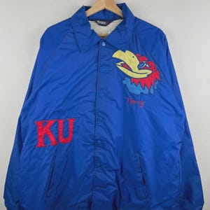 May include: A blue nylon jacket with a button-up front and a collared neckline. The jacket features the letters "KU" in red on the front and a colorful bird emblem with the name "Nancy" embroidered below it. The jacket is hanging on a wooden hanger.