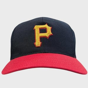 Puede incluir: Una gorra de béisbol con una corona de color azul marino oscuro y una visera de color rojo brillante. La parte frontal de la gorra presenta una letra P grande bordada en una fuente amarilla llamativa con un borde rojo fino, centrada.