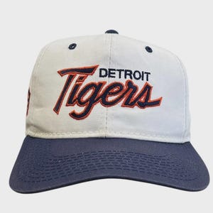 Puede incluir: Gorra de béisbol con corona blanca y visera azul marino. La parte delantera de la gorra presenta el texto "DETROIT Tigers" en una fuente estilizada con detalles en azul marino y naranja. La gorra tiene una visera curva.