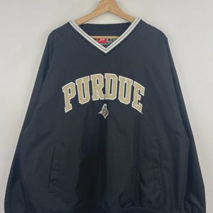 May include: Black Purdue pullover windbreaker with a V-neck and gold and white detailing. The word "PURDUE" is arched across the chest in gold lettering. Features a small logo below the text and two front pockets.