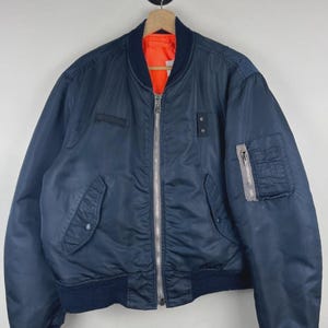 May include: A navy blue bomber jacket featuring a bright orange interior lining, a full-length silver metal front zip, ribbed knit cuffs and hem, two front snap-button pockets, and a zipped utility pocket on the left sleeve with a small tab.