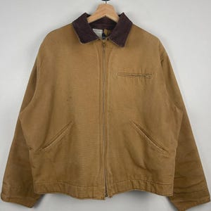 Op de afbeelding: Een bruine werkjas met een bruine corduroy kraag, een volledige ritssluiting en twee voorzakken. De jas hangt aan een houten hanger tegen een witte achtergrond.