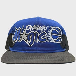 Może przedstawiać: Królewsko-niebieska czapka z daszkiem z wyhaftowanym napisem Orlando Magic białymi i niebieskimi literami. Wzór zawiera srebrną gwiazdę, grafikę piłki do koszykówki i małe białe gwiazdki. Czapka ma czarny daszek i czarne panele boczne.