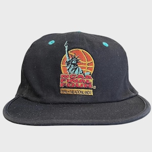 Könnte beinhalten: Eine schwarze Baseballkappe mit einem gestickten Logo der Freiheitsstatue über einer Basketballgrafik. Der Text lautet NCAA FINAL FOUR 1996 MEADOWLANDS in Rot und Gelb. Die Kappe hat kleine türkisfarbene Akzente auf den Kronenpaneelen.