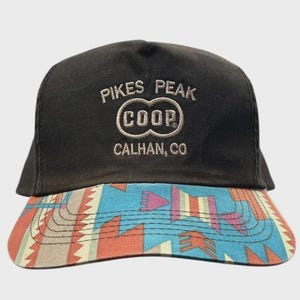 Puede incluir: Una gorra de béisbol negra con el texto bordado PIKES PEAK COOP CALHAN, CO en hilo color canela claro. La visera presenta un vibrante patrón geométrico multicolor en tonos azul, naranja, rosa y crema con costuras horizontales.