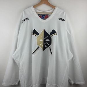 May include: A white long-sleeve V-neck jersey featuring a central graphic of a split gold and black goalie mask over two crossed hockey sticks. The text "SP" appears on the chest and inner collar, with "EFFIN" printed on the right shoulder.
