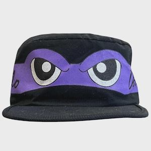 以下が含まれることがあります： 前面に幅の広い紫色の帯が入った黒いミリタリースタイルのキャップ。紫色の帯には、黒い瞳と白い虹彩を持つ、大きく様式化された漫画の目のグラフィックが描かれています。側面には黒い筆記体で「Don」という文字がプリントされています。