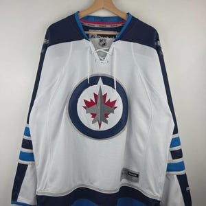 May include: A white Reebok Winnipeg Jets hockey jersey featuring a lace-up collar, navy blue shoulders, and a central circular logo with a silver jet and red maple leaf. The sleeves have navy and light blue stripes, with an NHL shield at the neck.