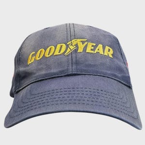 May include: A navy blue baseball cap featuring the word GOODYEAR embroidered in yellow thread across the front panels. The cap has a structured crown, a curved brim with visible stitching, and a classic six-panel design with a button on top.