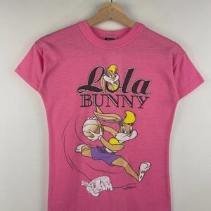 May include: A pink t-shirt featuring Lola Bunny from Space Jam. The graphic shows Lola dribbling a basketball, with the words "Lola Bunny" above and "Space Jam" below. The shirt is hanging on a wooden hanger.