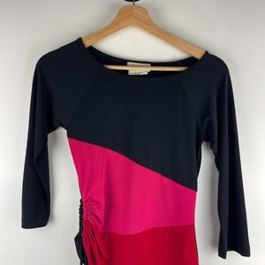 May include: A black, long-sleeved top with a color-blocked design. The top features a diagonal stripe of hot pink and a red stripe. The side has a ruched detail with a drawstring.