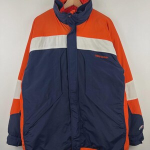 Peut inclure: Veste orange, blanche et bleu marine avec le mot "Illinois" brodé sur la poitrine. La veste a un col montant et une fermeture éclair sur toute la longueur. Une bande blanche traverse la poitrine et les manches.