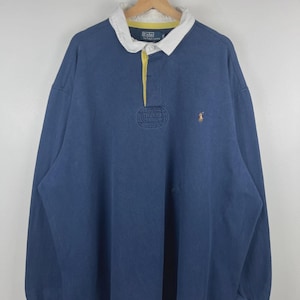 May include: A long-sleeve navy blue rugby shirt featuring a white collar with a yellow inner lining, a button placket, and an embroidered gold-colored logo on the chest. The shirt includes a Polo by Ralph Lauren label on the inner neck area.
