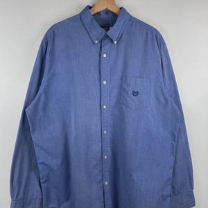 May include: A long-sleeve button-down shirt in a solid medium blue color. The shirt features a button-down collar, a front button placket, and a single chest pocket on the left side with a small, dark blue embroidered logo centered on the pocket.