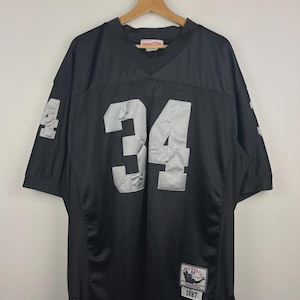 Pode incluir: Camisola de futebol preta com o número 34 prateado na frente e nas mangas. A camisola tem decote em V e uma etiqueta Mitchell & Ness throwback. A camisola está pendurada num cabide de madeira.