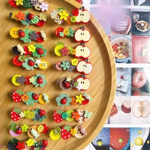 Può includere: Una collezione di fermagli per capelli decorativi con vari disegni, tra cui coccinelle, fiori, mele e fragole. I fermagli presentano bottoni colorati e dettagli in tessuto, disposti su un vassoio di legno.