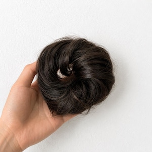 Könnte beinhalten: Ein dunkelbraunes Haarknoten-Haarteil mit einer kreisförmigen, voluminösen Form und geschichteten Strähnen. Das Haarteil hat eine natürliche Textur und eine abgerundete, leicht zerzauste Optik, die dazu dient, einen vollen Dutt zu stylen.