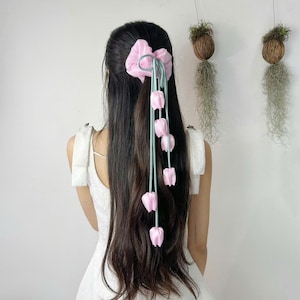 May include: A pink scrunchie with a bow and light blue ribbon strands, each with a pink tulip. The hair accessory is set against long, dark hair and a white dress. The background includes hanging plant decorations.