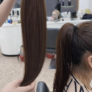 Op de afbeelding: Een bruine haarextensie, bevestigd aan een zwarte elastiek, wordt omhoog gehouden. De afbeelding toont de lengte en kleur van het haarstuk. Een persoon met bruin haar in een paardenstaart is ook zichtbaar.