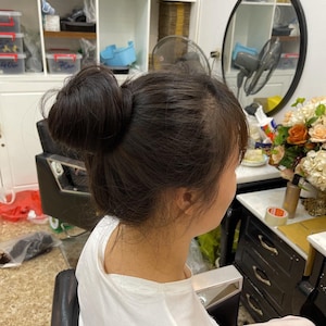 Könnte beinhalten: Eine dunkelbraune Haarknoten-Frisur mit losen Strähnen, die das Gesicht umrahmen, kombiniert mit einem schlichten weißen T-Shirt mit kurzen Ärmeln. Das Haar ist am Oberkopf zu einem runden, strukturierten Knoten zusammengefasst.