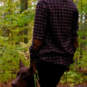 Könnte beinhalten: Eine Person trägt ein weinrotes kariertes Hemd mit braunen Ellbogenflicken und schwarzer Hose und steht in einem Wald mit einem braunen Hund an der Leine. Der Hund hat ein silbernes Kettenhalsband.