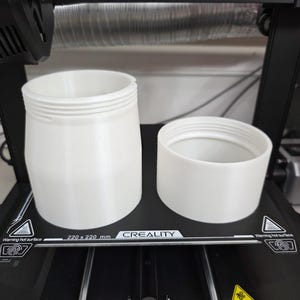 May include: Two white, cylindrical 3D-printed containers with threaded openings. One container is taller and wider than the other. The containers are on a black surface with a warning label that reads "Warning hot surface".