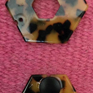 May include: Two hexagonal tortoise shell patterned plastic pieces with a hole in the center. The top piece has a smaller hexagonal hole in the center. The bottom piece has a black button in the center.