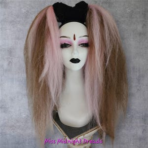Könnte beinhalten: Ein Schaufensterpuppenkopf mit schwarzem Stirnband und Schleife, gestylt mit langen, hellbraunen und rosa Dreadlocks. Die Schaufensterpuppe hat auffälliges Make-up, darunter schwarzer Lippenstift und ein rotes Bindi. Der Text "Miss Midnight Dreads" ist unten sichtbar.