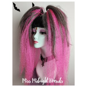 May include: Pink and black synthetic dreadlocks with two black bat hair clips. The dreadlocks are styled in two high ponytails. The dreadlocks are on a mannequin head.