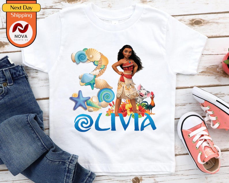 May include: White t-shirt with a colourful design featuring the name "Olivia" in blue, a cartoon character, and the number "2" decorated with seashells. The shirt also has a "Next Day Shipping" logo.