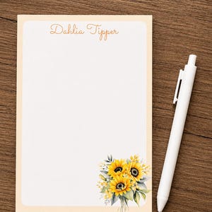 May include: A rectangular notepad with a light peach border and white center, featuring the text "Dahlia Tipper" at the top and a cluster of yellow sunflowers with green leaves in the bottom right corner, accompanied by a plain white ballpoint pen.